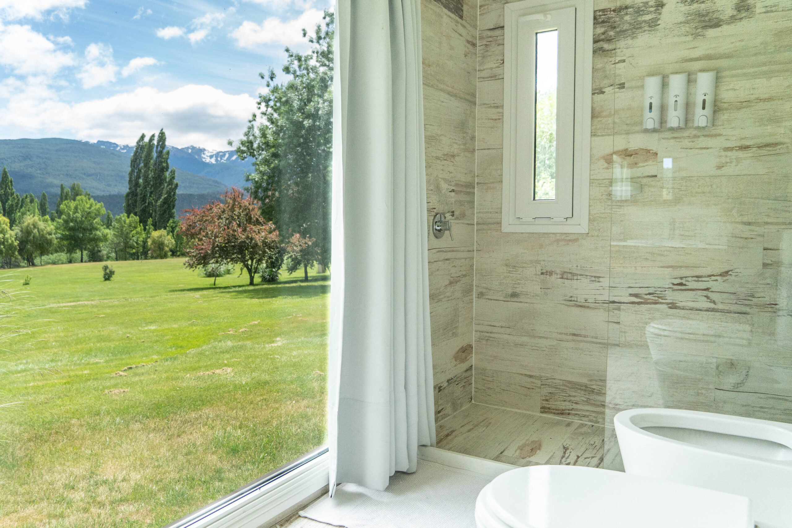 Baño moderno en La Aguada Suite con ducha, revestimiento claro y ventana con vista a montañas en El Bolsón