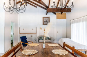 Comedor con mesa de madera y sillas rústicas, candelabros colgantes y techo con vigas, integrado a una sala luminosa con sillones.